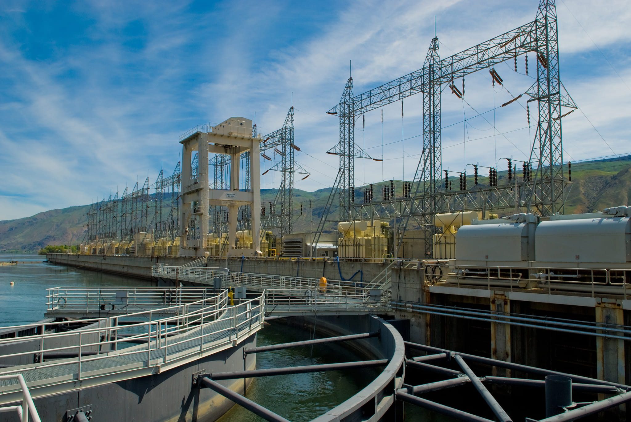 Hydroelectric dam switchyard with concrete gantries, transformers, MV/HV transmission towers and walkways along the waterway