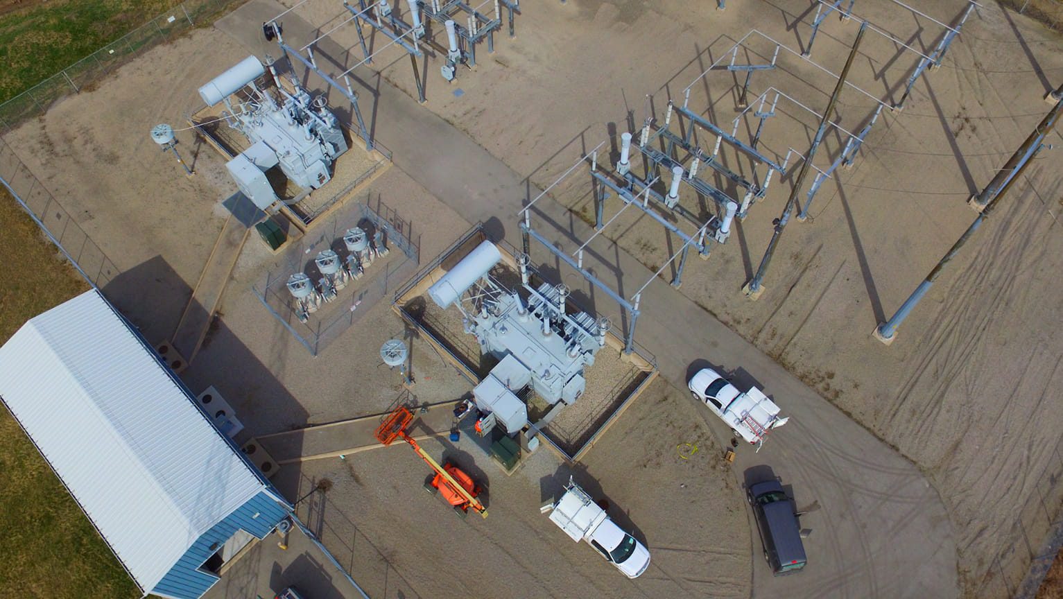 Aerial view of fenced substation with large transformers, switchgear, overhead lines, service trucks and an orange boom lift