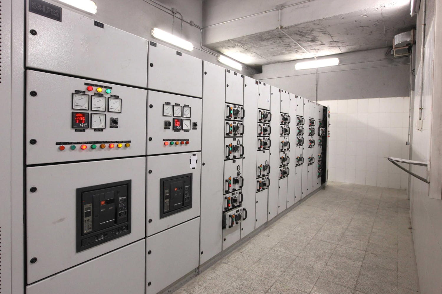 Metal electrical switchgear cabinets with analog meters, indicator LEDs and breakers lining a service room corridor