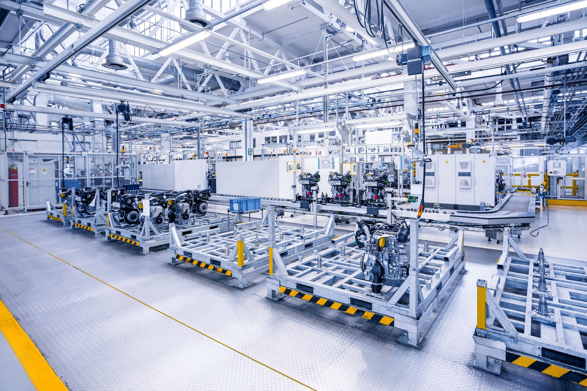 Bright industrial assembly line with metal frames, engines on skids, overhead cable trays, conveyors and plant lighting