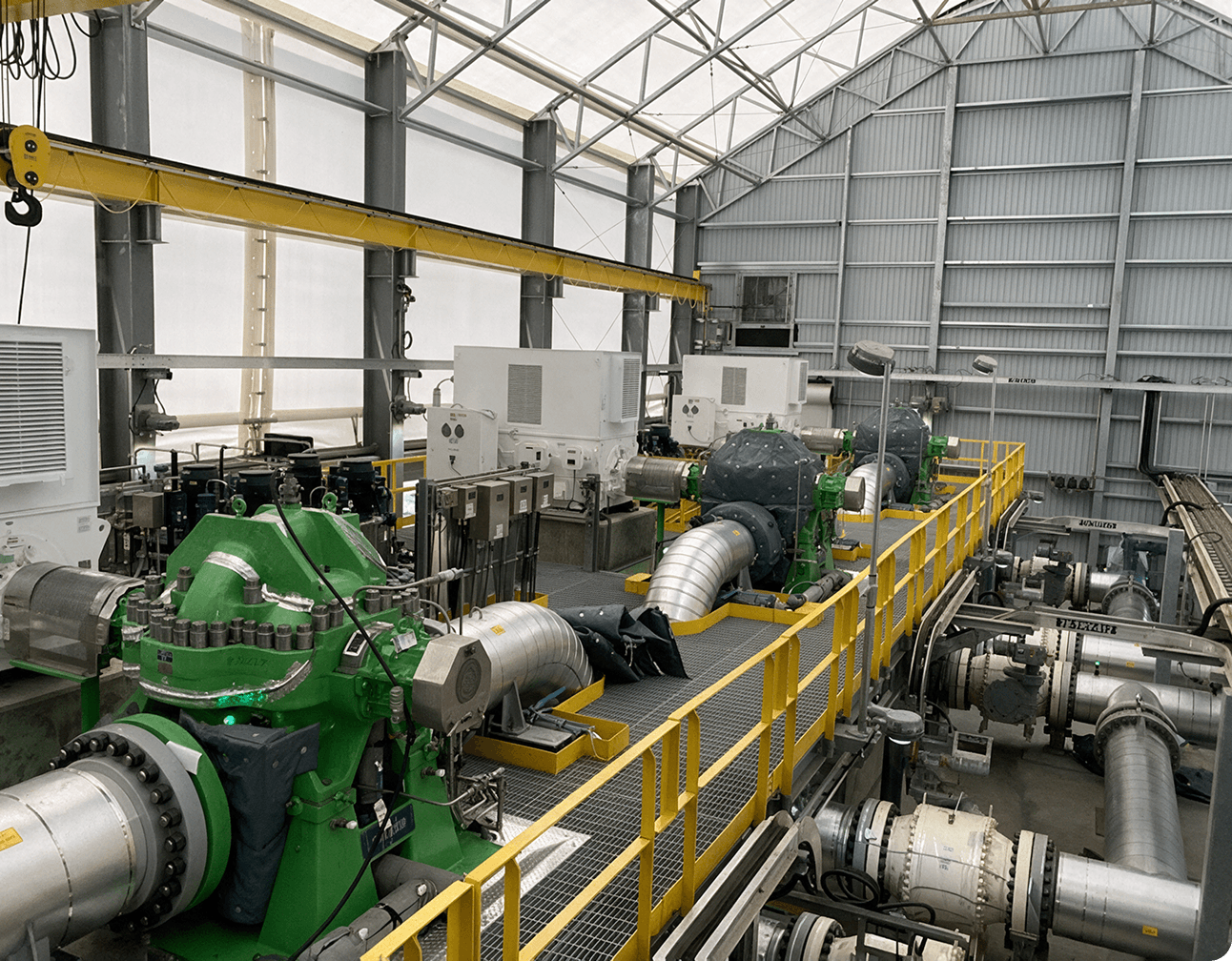 Industrial pump room with green pumps, large stainless pipes and yellow-railed grated walkway under an overhead crane