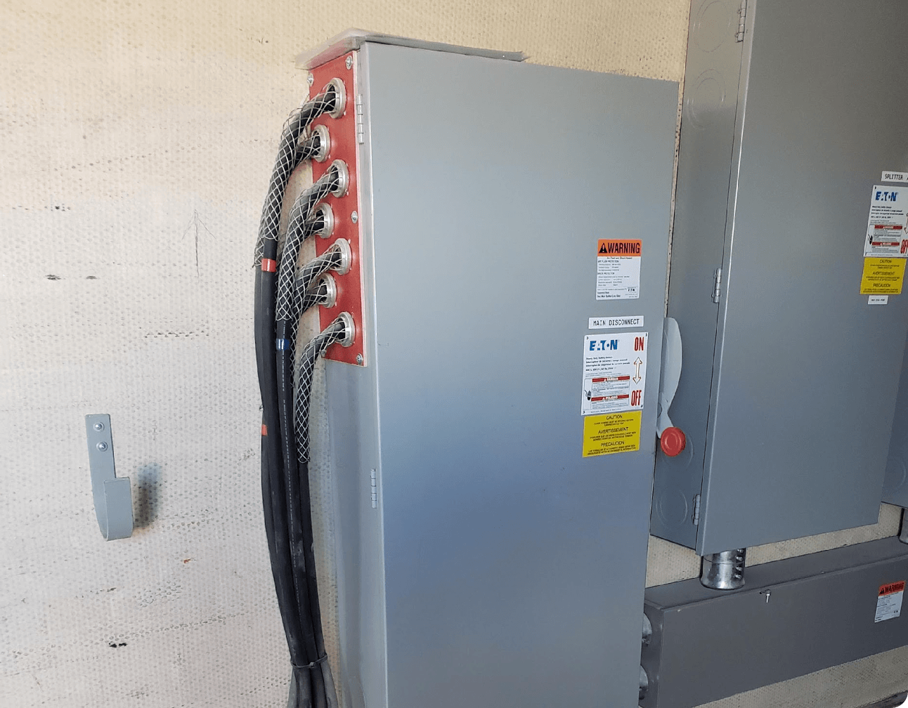 Metal electrical disconnect cabinet with six armored power cables entering a red feed plate and warning stickers on the door