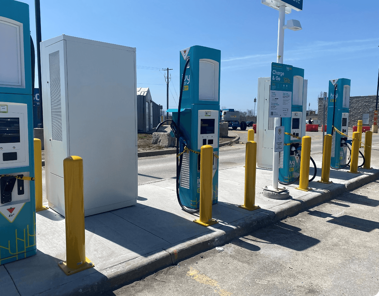 Dual-head electric vehicle fast chargers on a concrete island with yellow bollards and a white service cabinet