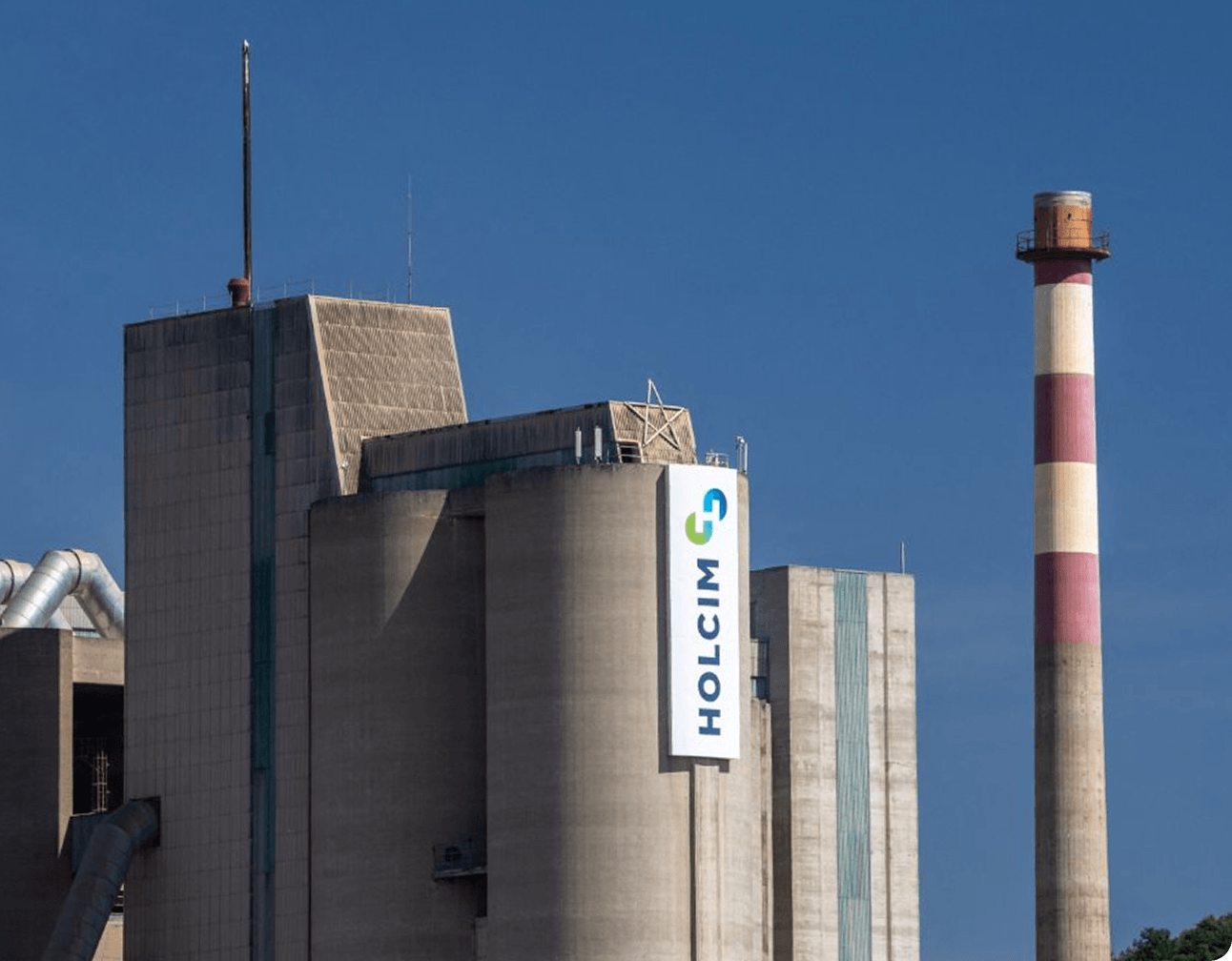 Large concrete industrial plant silos and a striped smokestack under blue sky with ventilation ducts and rooftop equipment