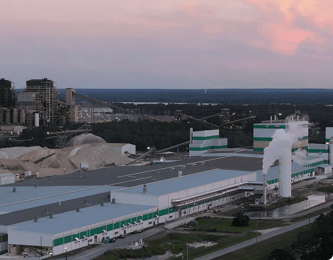 Aggregates processing plant at dusk with large warehouses, conveyors, material piles and steam venting from exhaust stacks