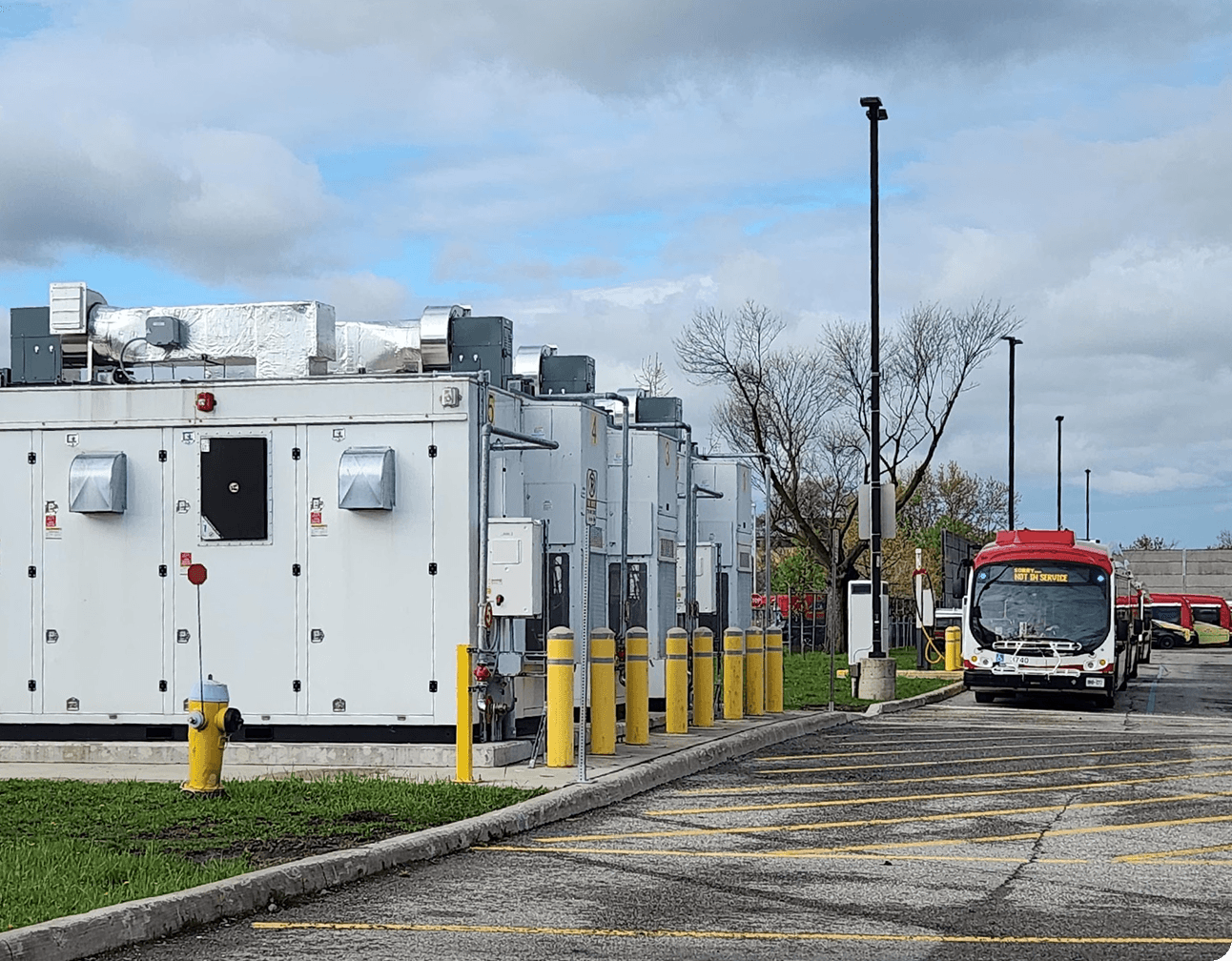 Electric bus charging site with large white power cabinets, rooftop vents, yellow bollards and a red municipal transit bus