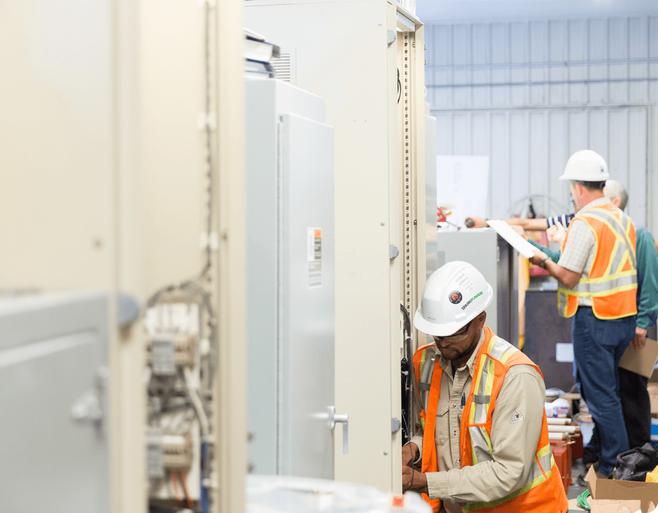 Electricians in hard hats and orange vests working on PLC control cabinets and wiring inside a roastery production area