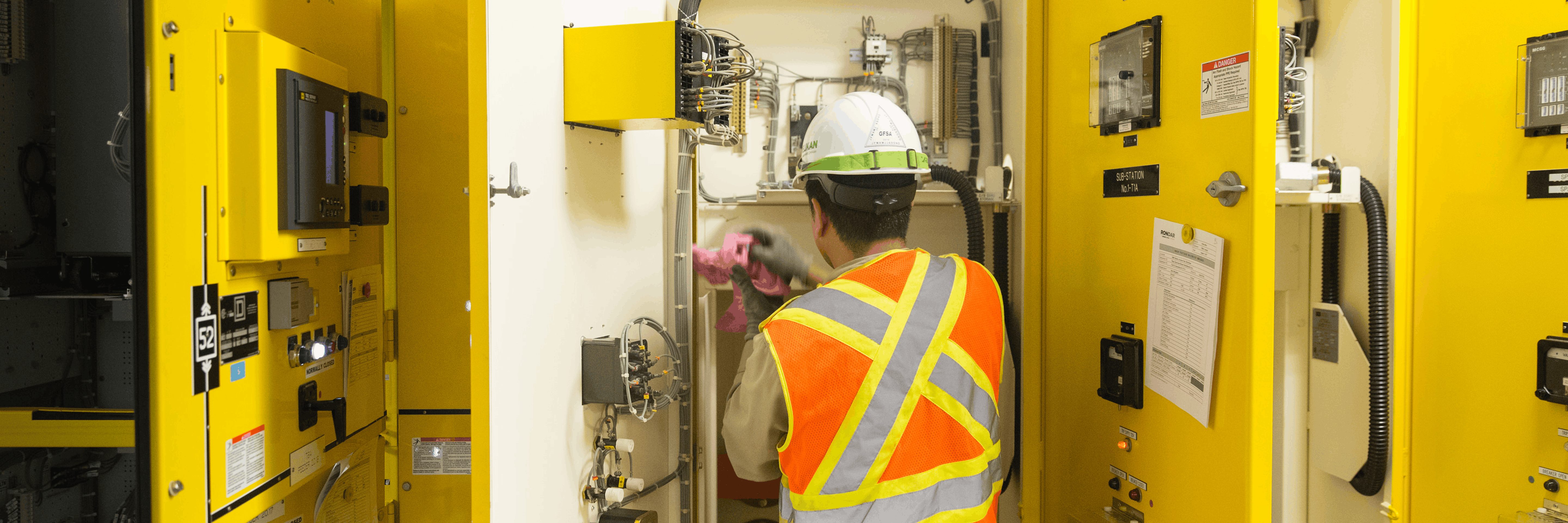 Worker in safety vest and hard hat inspecting and cleaning yellow electrical switchgear panels in a utility room