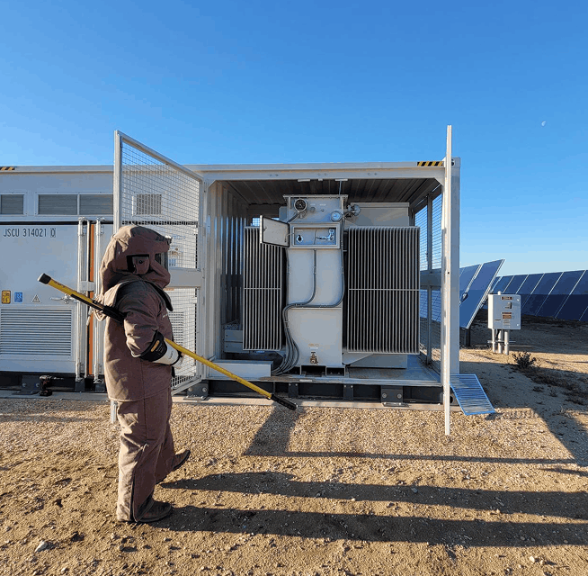 Technician in cold-weather protective suit using a grounding stick to inspect an outdoor transformer at a solar site