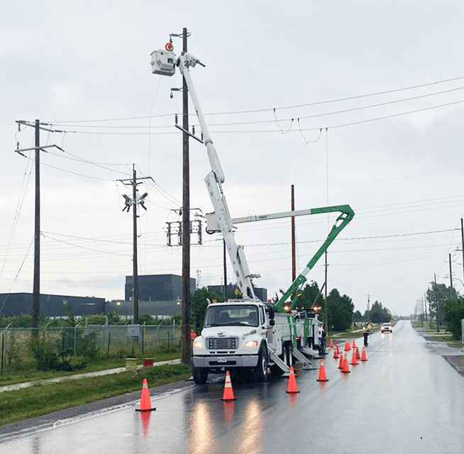 Bucket trucks and linemen repairing overhead power lines on a rainy road with traffic cones and safety perimeter