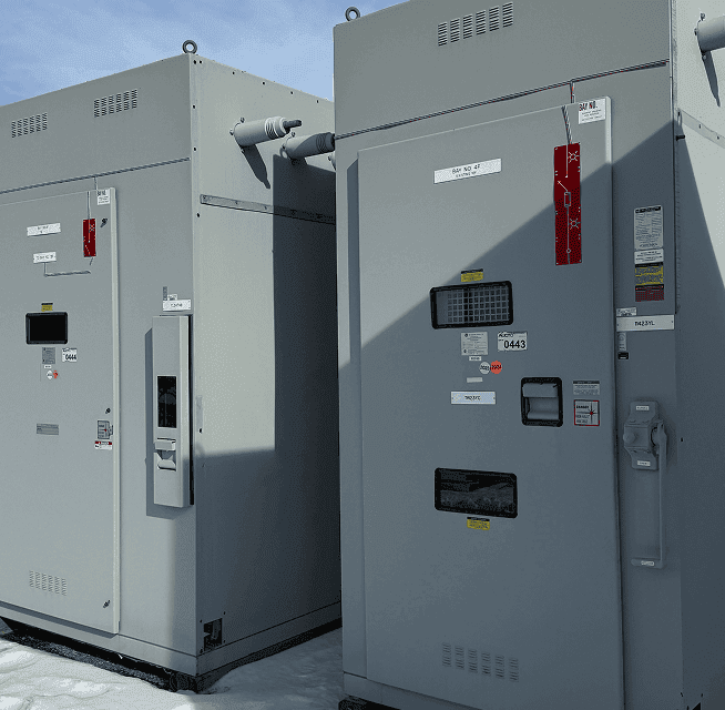 Two large gray electrical equipment cabinets outdoors on snow-covered ground with access panels and warning labels visible