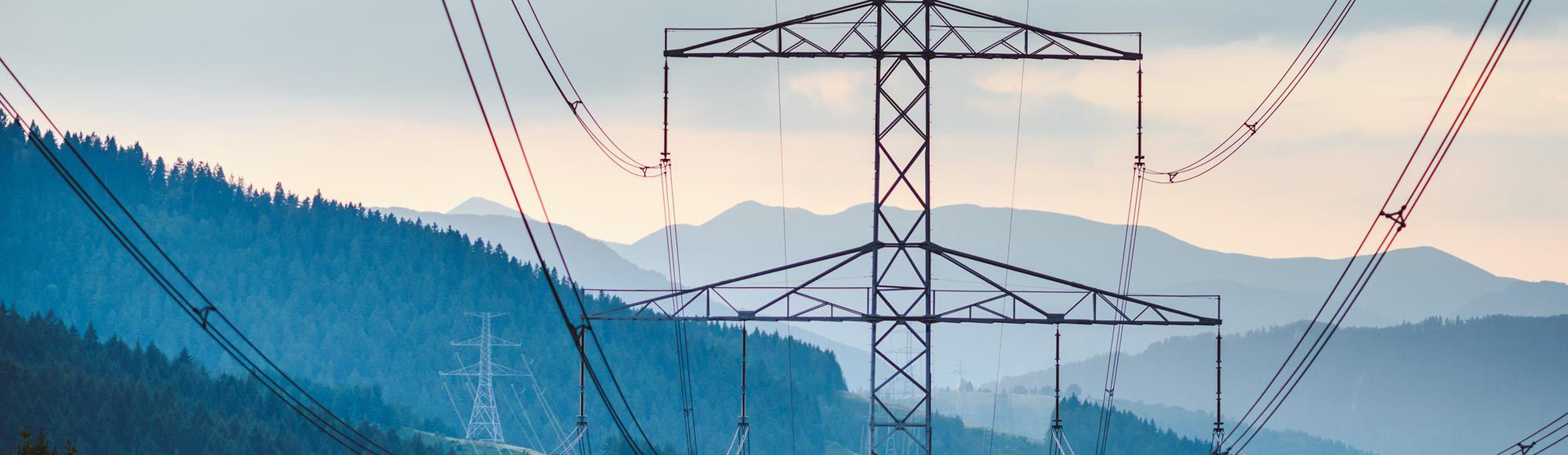 High-voltage transmission towers and power lines crossing forested hills with layered mountain silhouettes at dusk