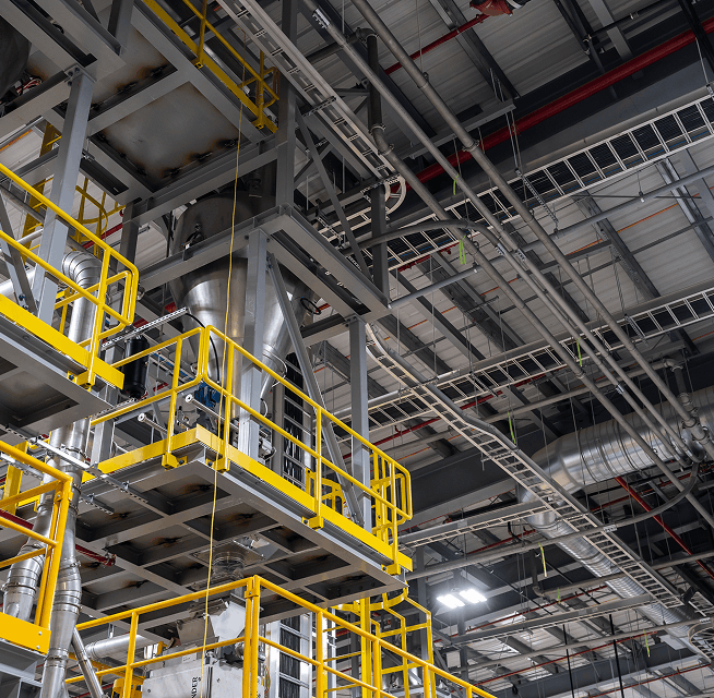 Large stainless hoppers and conveyors with yellow railings under overhead piping and cable trays in an industrial plant