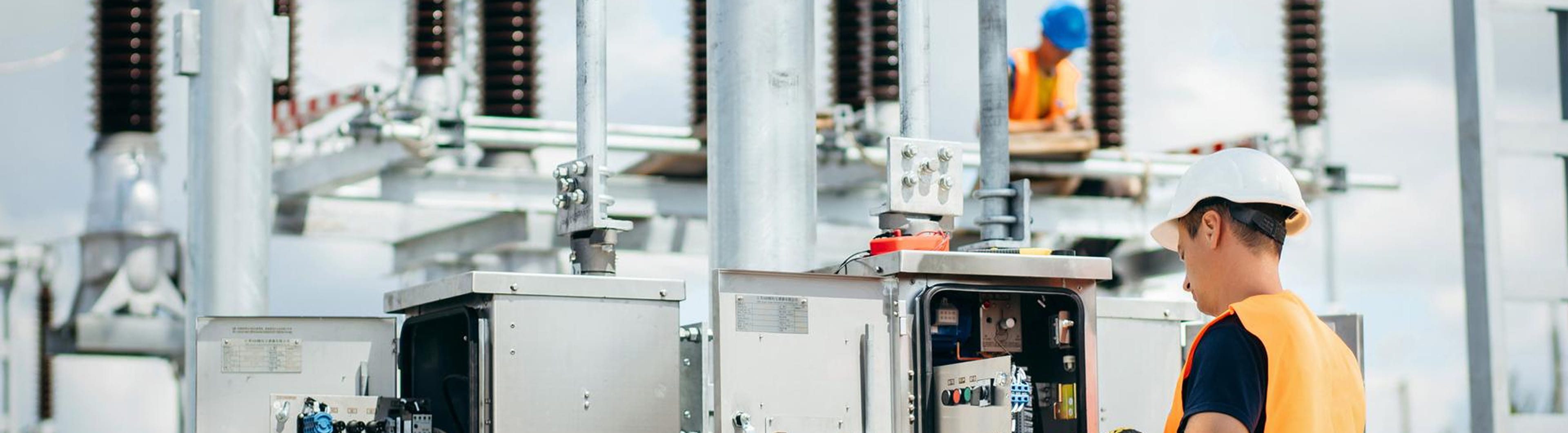 Worker in hard hat and reflective vest inspecting substation control cabinet with exposed wiring and metal support structures