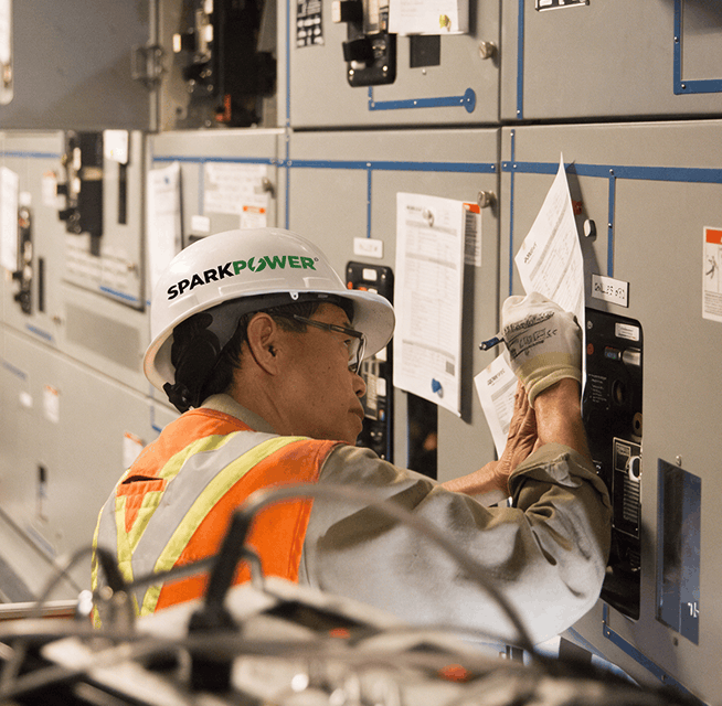 SparkPower technician in safety vest and hard hat inspecting and recording settings on electrical switchgear panels