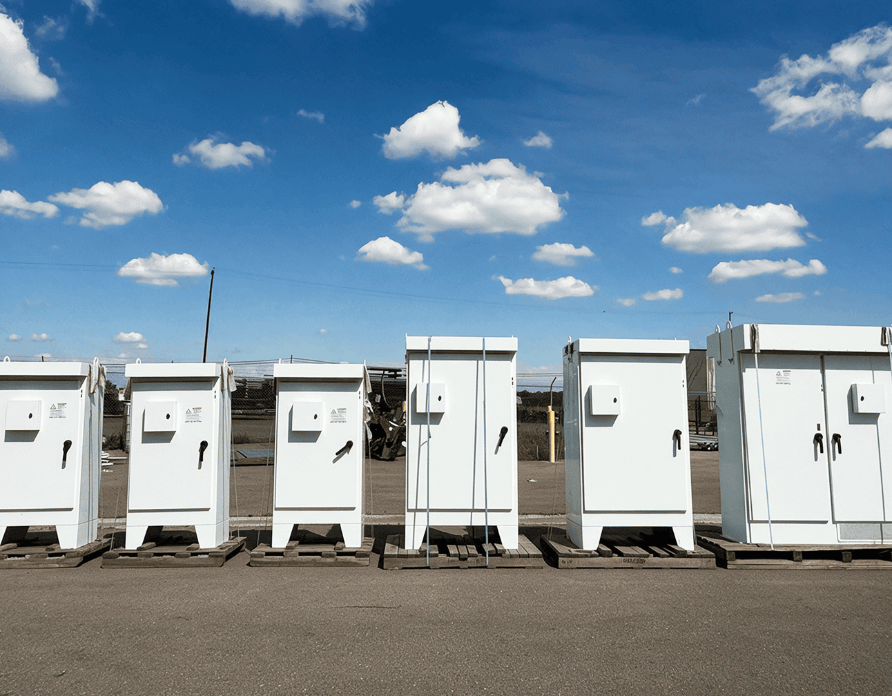 White outdoor electrical control cabinets and motor control panels on wooden pallets in a fenced yard under clear skies