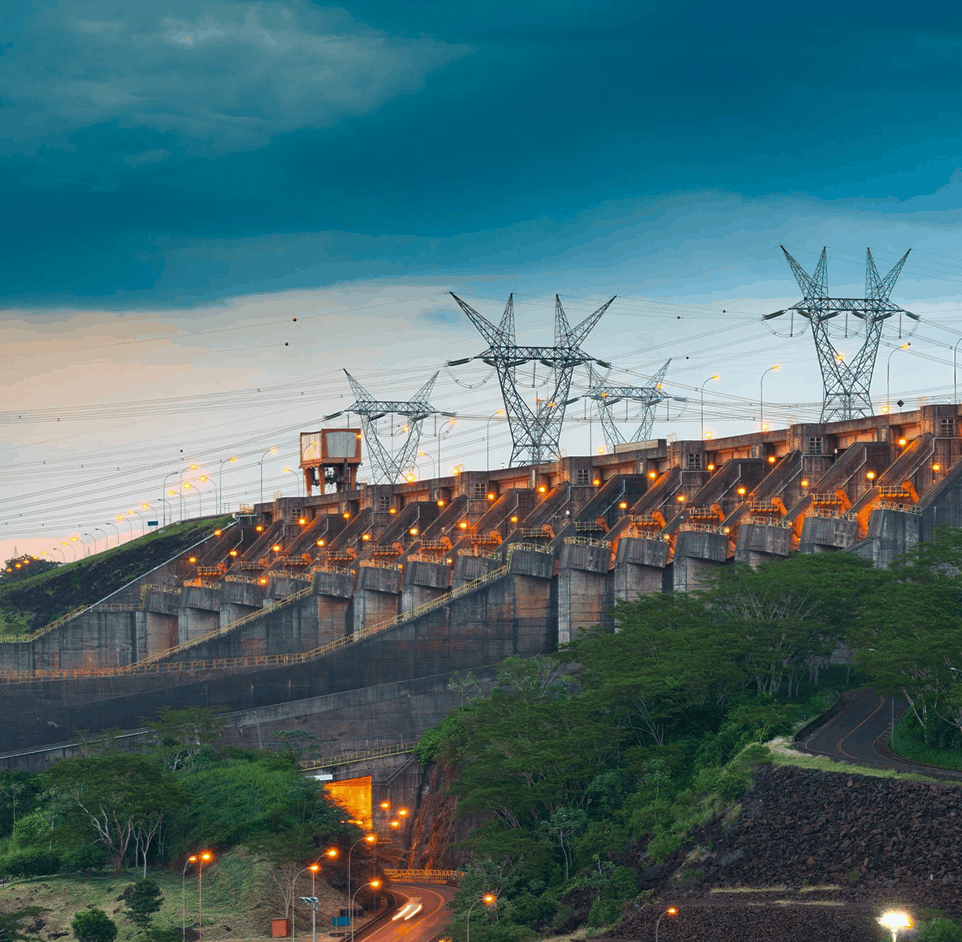 Hydroelectric dam with multiple gated spillways, lit walkways and transmission towers carrying high-voltage lines at dusk