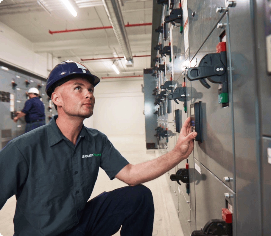 SparkPower technician kneeling and inspecting industrial electrical switchgear panels and control handles in a utility room