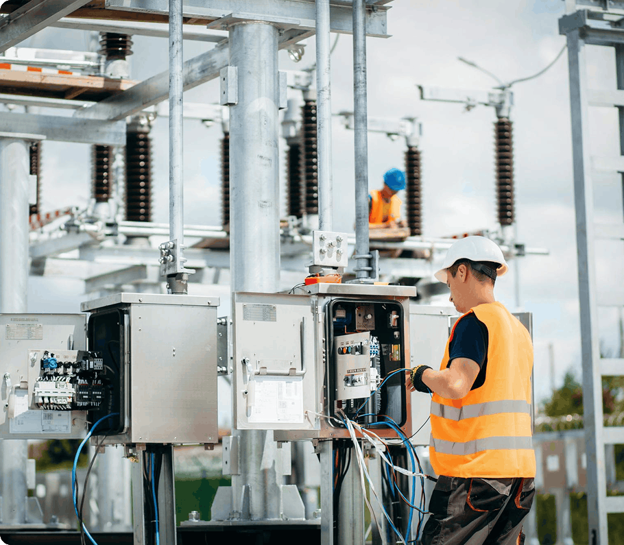 Technician in hard hat and safety vest wiring cables into outdoor control cabinets at an electrical substation