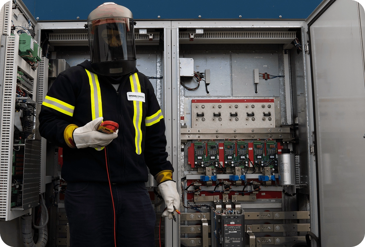 Technician in arc-flash suit and gloves holding test leads inspecting an open electrical control cabinet with circuit boards
