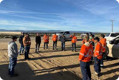 Field crew in orange safety gear and hard hats assembled for a morning safety meeting beside company trucks and power lines