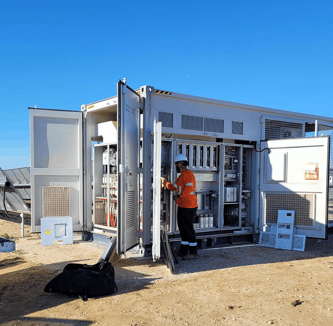 Technician in orange safety gear and hard hat working inside open electrical switchgear cabinets at a solar site