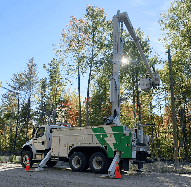Bucket truck with extended articulating boom and outriggers parked near a transmission pole in wooded area