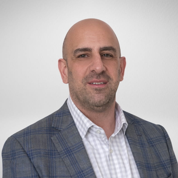 Professional headshot of Ali Assaf in a checked gray blazer and white dress shirt on a neutral light-gray background