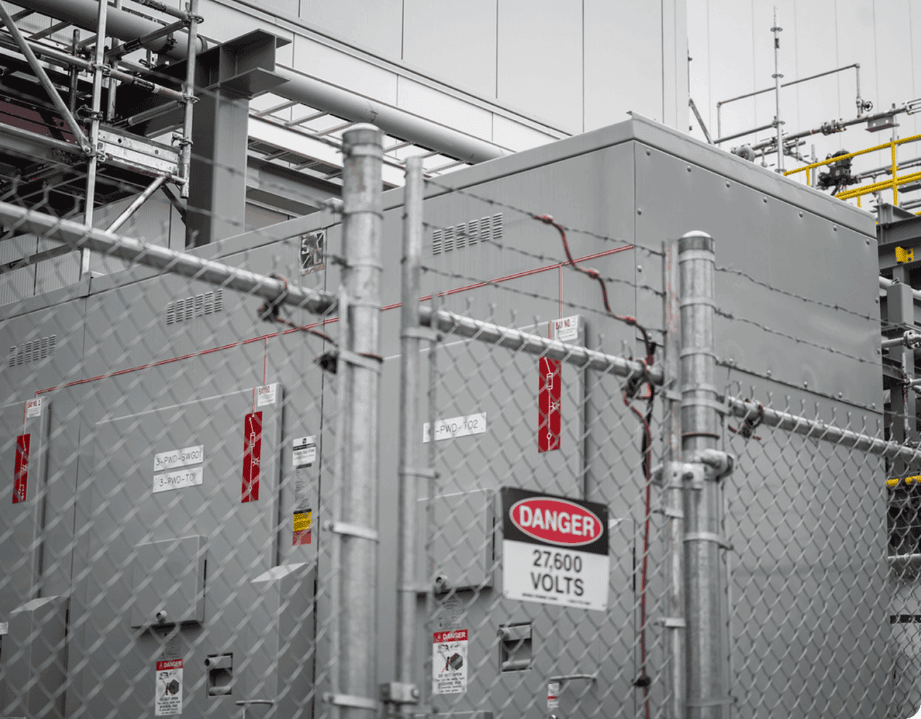 Outdoor industrial electrical transformer enclosure behind chain-link fence with warning signs and high-voltage cabling