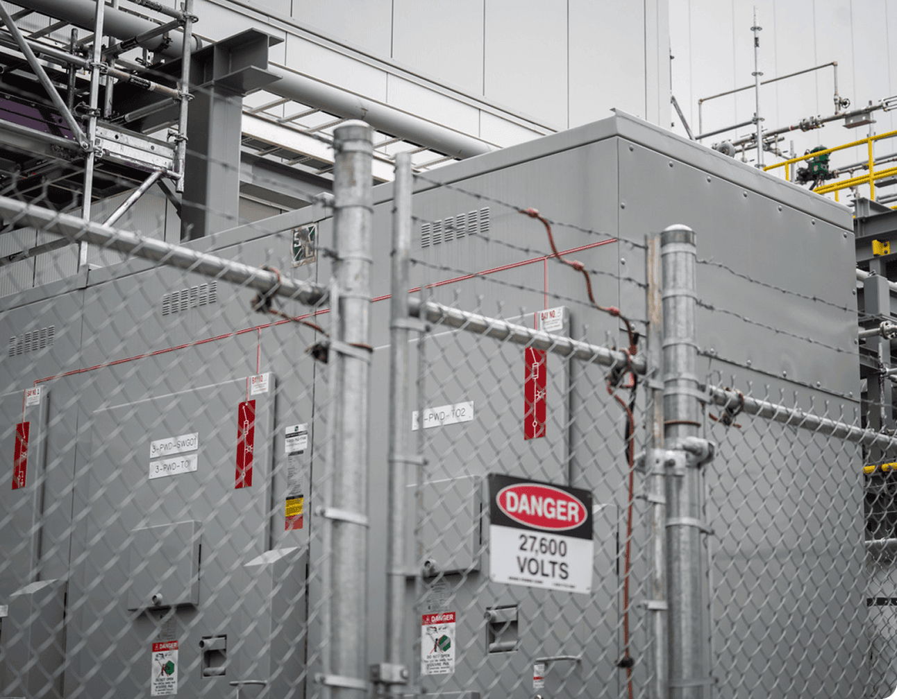 Fenced outdoor electrical transformer enclosure with high-voltage cabinets, warning signs and industrial piping