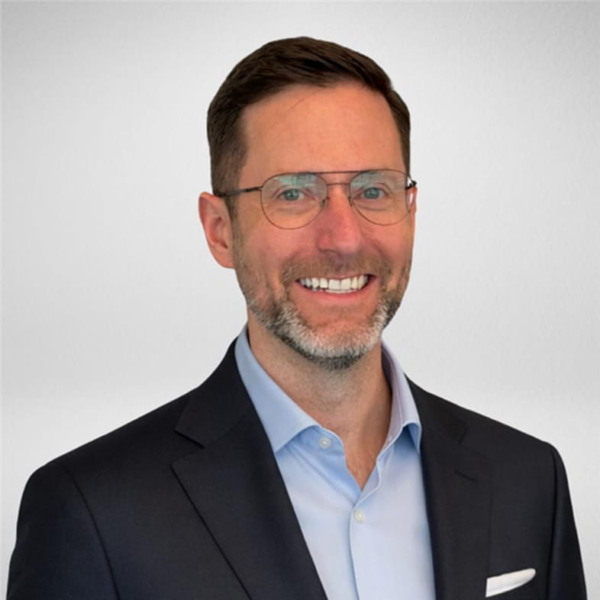 Smiling male professional with glasses in a navy suit jacket and light blue dress shirt on a neutral gray backdrop