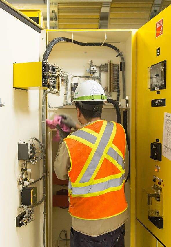 Field technician in hard hat and Hi‑Vis vest performing commissioning and testing inside open electrical switchgear cabinet