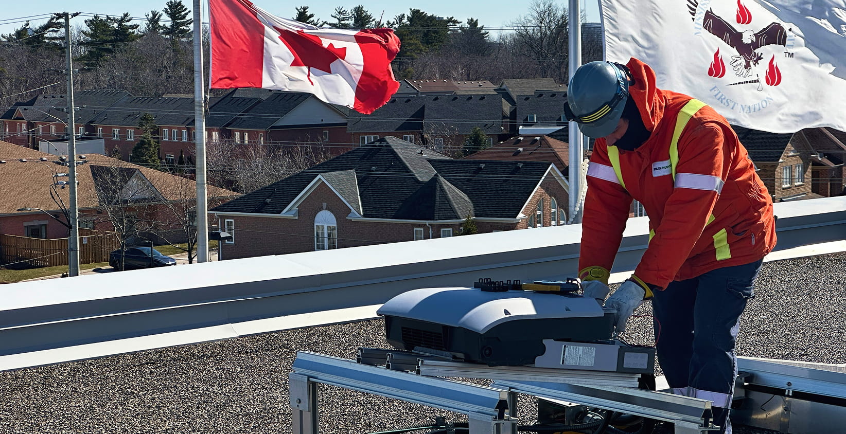 Technician in orange safety jacket and hard hat inspecting rooftop HVAC unit with Canadian flag behind residential homes