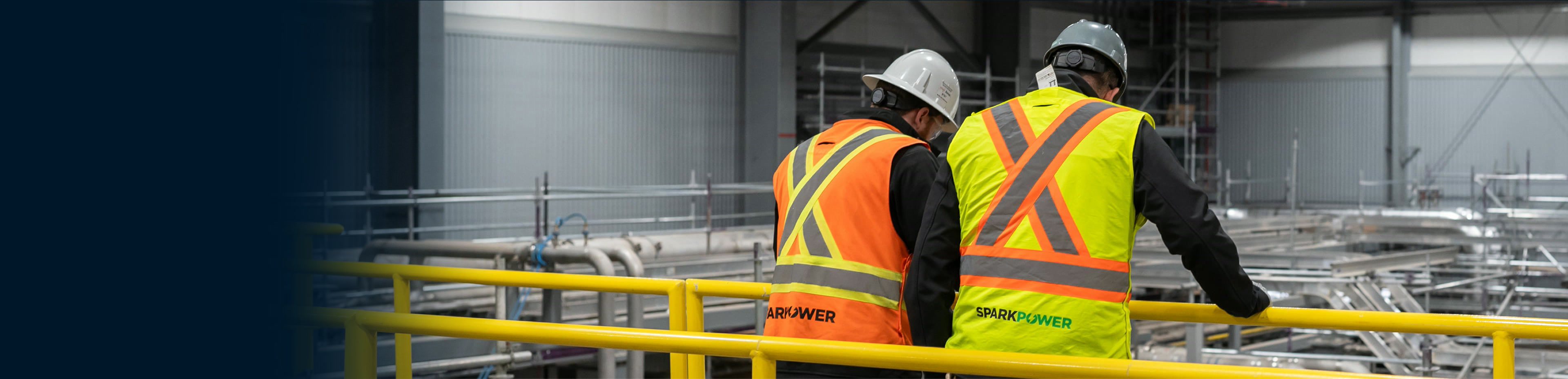 Two workers in hard hats and high-visibility vests inspect industrial piping and metal framework from a yellow safety railing