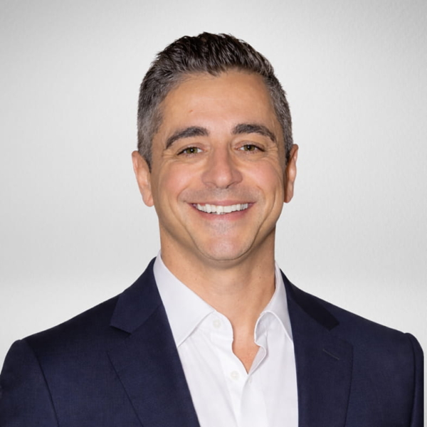 Smiling professional man in a dark suit jacket and white shirt against a neutral studio background, headshot for team profile