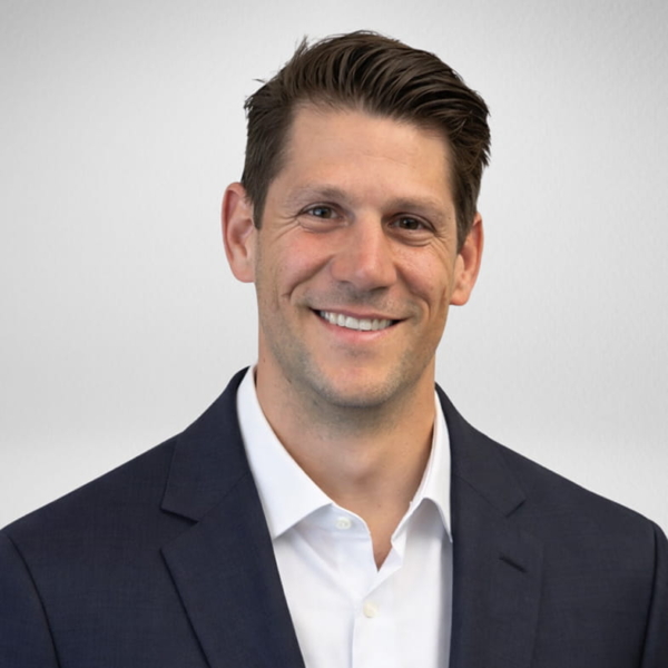 Male professional portrait wearing a navy suit jacket and white shirt, smiling against a gray backdrop