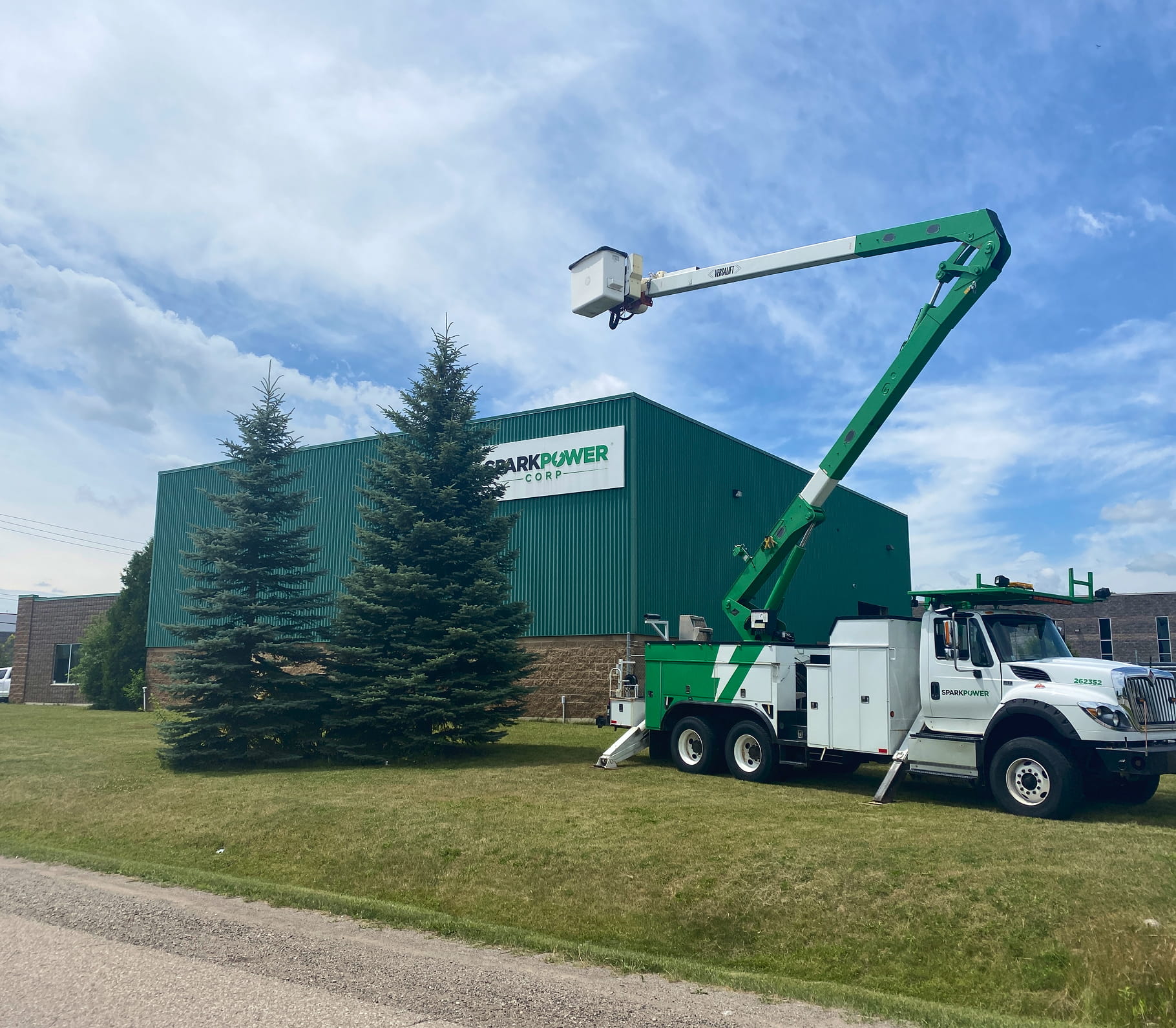 White and green bucket truck extended beside a green SparkPower building with evergreen trees and blue sky