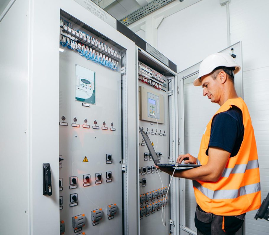 Electrician in hard hat and high-visibility vest using a laptop to test and control panels inside an electrical cabinet