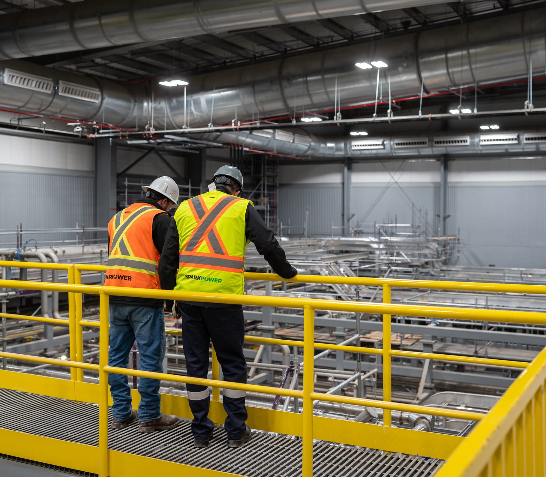 Two workers in safety vests and helmets observe large ductwork and piping from a yellow railing inside an industrial facility